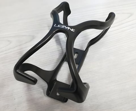 Lezyne Flow Cage in black, a plastic bottle cage with the brand logo, resting on a pale wooden surface.