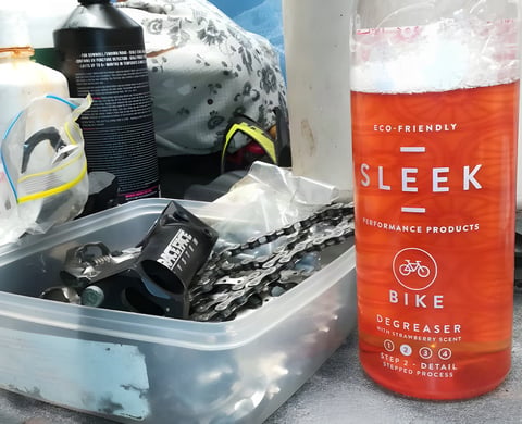Bottle of SLEEK Bike Degreaser with strawberry scent beside a tub of bike chains and parts.