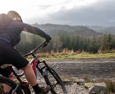 Wales360 rider on a full-suspension mountain bike on a stony forest track, with conifers and hills behind.