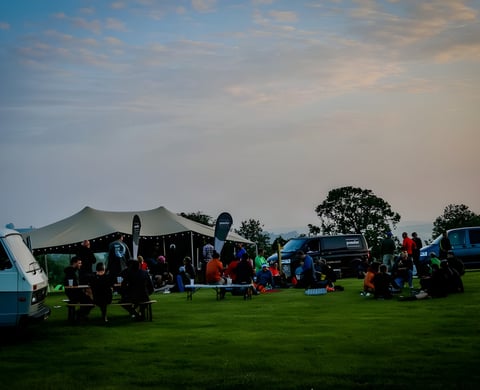 Riders sit on the grass at The Distance gravel event base camp beside a marquee and vans at dusk.