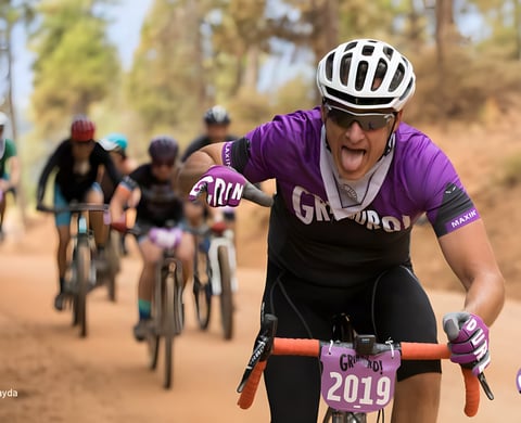 Grinduro rider in a purple jersey sticks out their tongue on a dusty trail, with a 2019 number board on the bars.