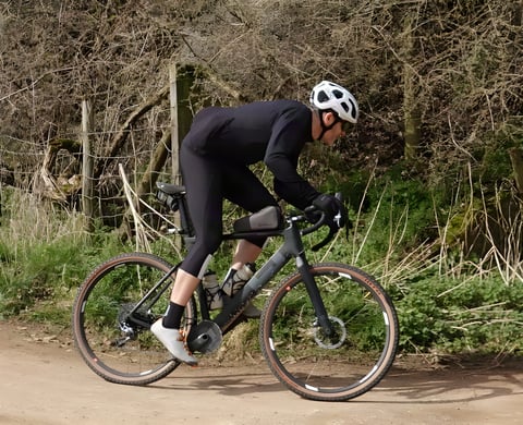 Rider wearing the 7Mesh Mission Jersey in black on a gravel bike on a dirt track beside a hedge.