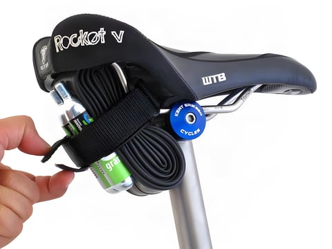 Backcountry Research Race Strap holding a spare tube and CO2 cartridge under a WTB saddle, a hand pulling the Velcro tab.
