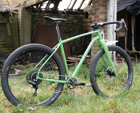 Green Mason InSearchOf steel bike with 29er tyres, drop bars and front rack, parked on grass outside a ruined barn.