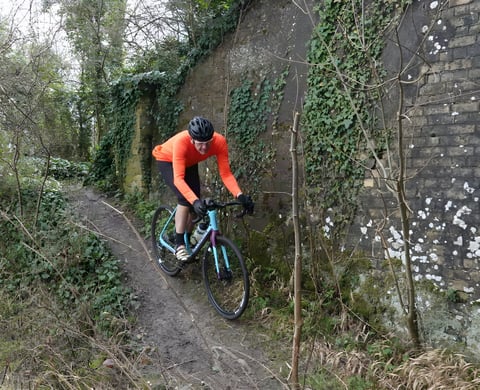Rider in the Very Orange Galibier Roubaix Long Sleeve Jersey descends a muddy track past an ivy-covered stone wall.