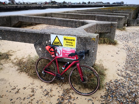 Sonder Camino AL leaning on a concrete sea defence by a beach, beside a 'Beware strong currents' sign.