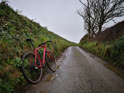 Sonder Camino AL leaning on a grassy bank beside a wet, puddled gravel lane between high hedges.