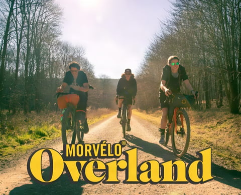 Three riders on a sunny gravel track with loaded bikes under the Morvélo Overland logo.