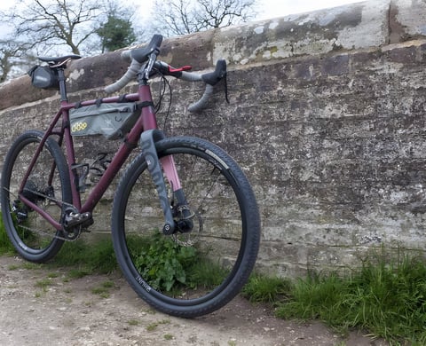 Rondo Ruut AL aluminium gravel bike in satin ruby with a frame bag, leaning against a stone wall.