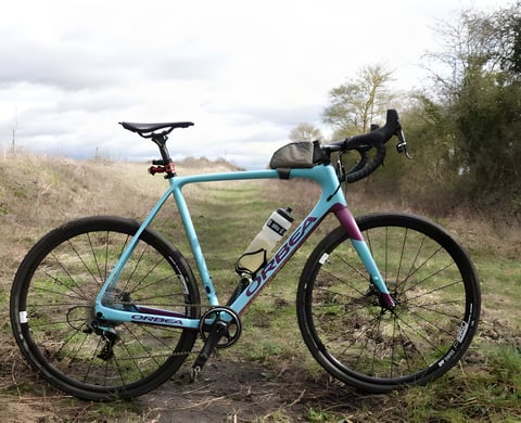 Orbea Terra M21-D 19 gravel bike in light blue and purple, parked on a grassy track.