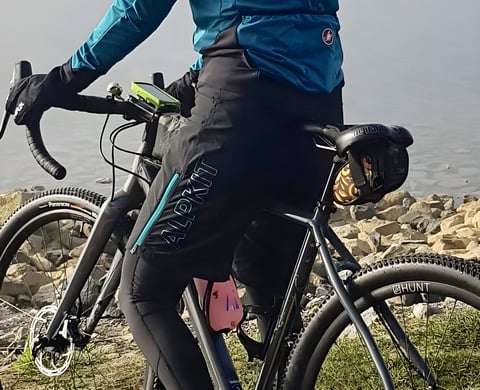 Rider in black Alpkit Floe women's MTB shorts with teal zip on a gravel bike beside water.