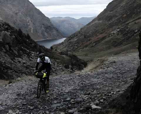 Rider with a bar bag climbs a rocky track above a reservoir, a promotional shot for the Further event in the Pyrenees.