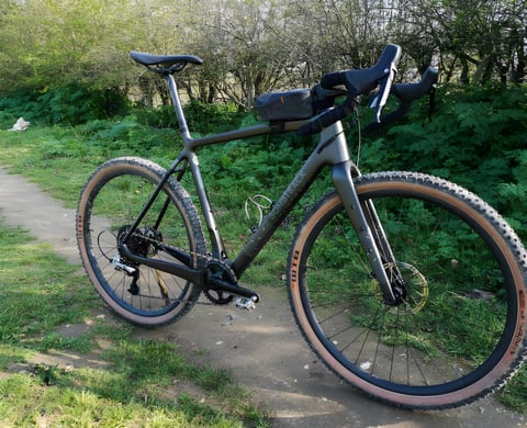 Bombtrack Hook EXT-C carbon gravel bike with a top-tube bag and tan-wall WTB Sendero tyres on a dirt track beside a hedge.