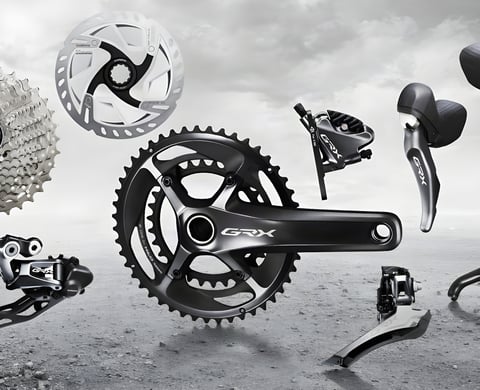Shimano GRX gravel groupset components, including crankset, derailleurs, brake caliper, rotor and shifter, on grey gravel.