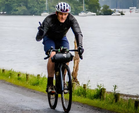 Rider in the black 7Mesh Oro jacket on a gravel bike with a bar bag, riding a wet lakeside road in the rain.