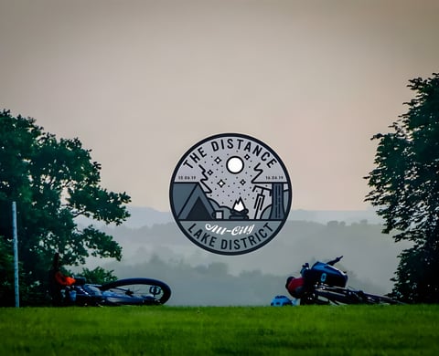 The Distance 2019 Lake District badge over two bikes lying on grass at dusk above a misty valley.