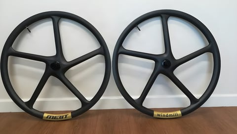 Pair of Merit Windmill five-spoke carbon gravel wheels with gold rim decals, leaning against a wall.