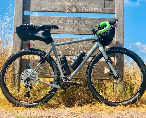 Axel Carion's OPEN WI.DE gravel bike with Ortlieb bags and WTB tyres, leaning on a wooden trail signpost in dry grass.