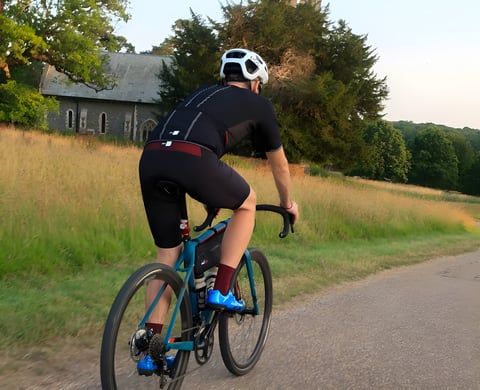 Rider in PEdALED Odyssey jersey and bibshorts on a teal gravel bike, passing a stone church on a quiet lane.