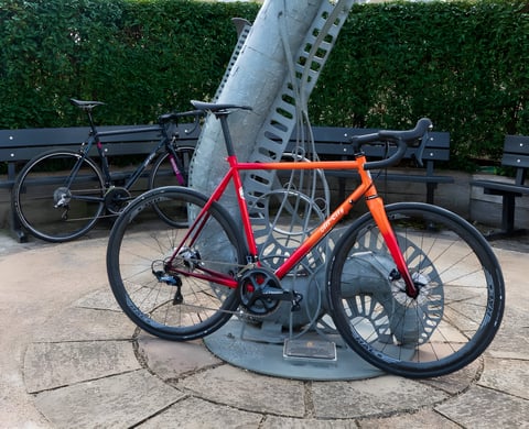 All City Zig Zag in the orange-red rum punch fade, with Halo wheels, leaning on a metal sculpture in a park.