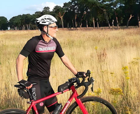 Rider in the Sundried Peloton jersey and shorts stands astride a red Pinnacle gravel bike in a field of tall grass.