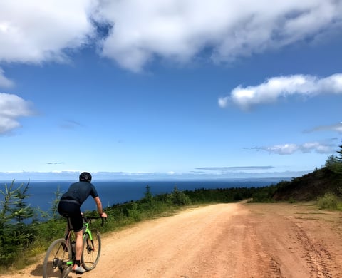 Rider on a green gravel bike climbs a sandy track above a deep blue sea on a summer gravel holiday.