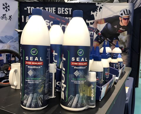 Bottles of Squirt SEAL tubeless tyre sealant with BeadBlock on a display table, a rider pictured on the banner behind.