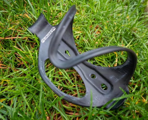 Arundel DTR/STR carbon bottle cage lying on grass, showing its side-entry shape and mounting holes.