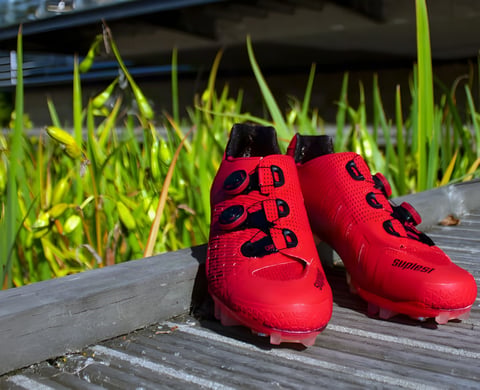 Pair of neon red Suplest Cross Country Pro BOA cycling shoes on wooden decking beside long grass.