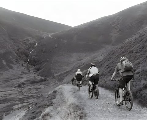 Three riders climb a gravel track up a steep-sided Lake District valley on the Low Key Lakes Weekender, in black and white.