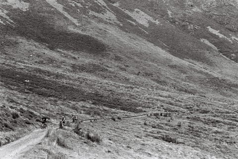 Three tiny riders on a track crossing a vast steep fellside, giving a sense of the Lake District's scale.