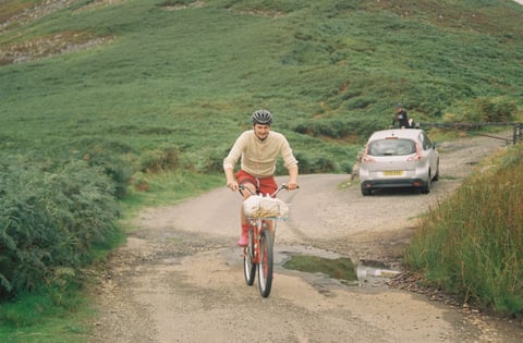 Rider on a red bike with a front basket rolls along a puddled gravel track below a bracken-covered fellside.