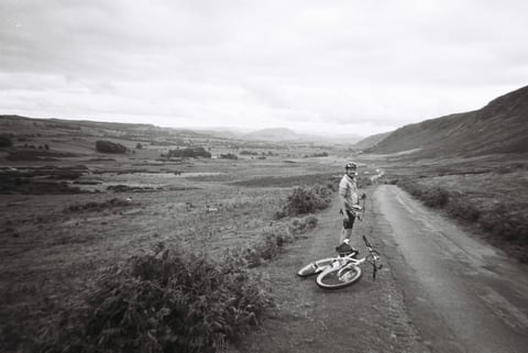 Rider stands beside a bike lying on the verge of a quiet road running through a wide Lake District valley.