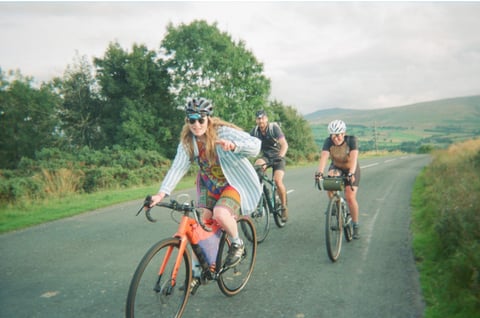 Three riders on a quiet Lake District lane, the front rider on an orange gravel bike pointing at the camera.