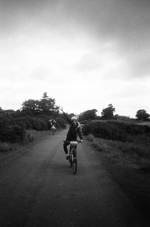 Rider raises an arm in celebration while riding along a quiet lane, another rider behind, in black and white.