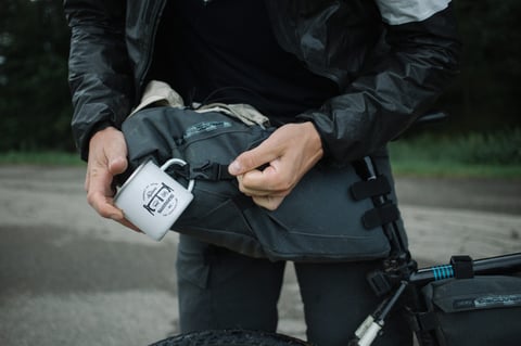 Rider fastens a Shimano enamel mug to a PRO Discover seat pack beside a loaded gravel bike.