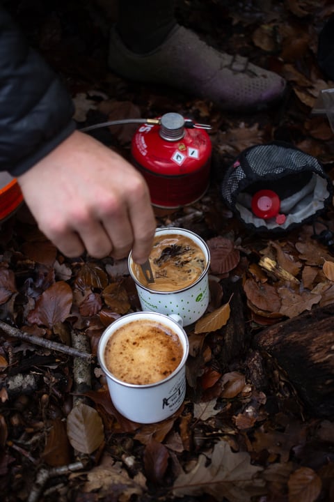 Hand stirs frothy coffee in an enamel mug beside a second mug and a red gas canister on forest leaves.