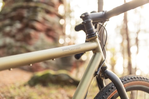 Tout Terrain logo on the top tube of a Vasco gravel bike, with stem, handlebar and fork in soft woodland light.