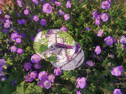 An Africap cycling cap in white, brown and green Ankara print lies among purple flowers.
