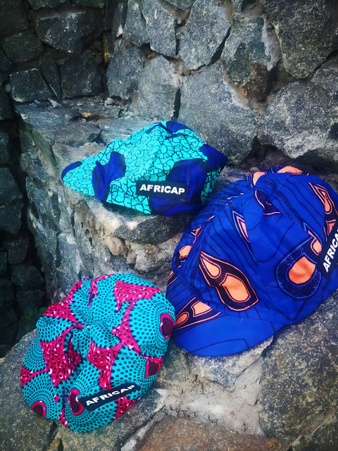 Three Africap cycling caps in bold blue, turquoise and pink Ankara fabric rest on a stone wall.