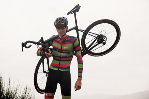 CX Syndicate rider in team kit shoulders a cyclocross bike against a misty sky.