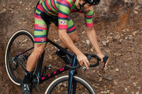 CX Syndicate rider in striped Stolen Goat team kit leans over the bars of a navy Vitus cyclocross bike on a leafy trail.