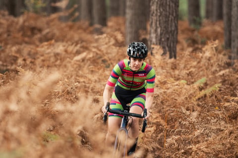 CX Syndicate rider in striped team kit rides through brown bracken in a pine forest.