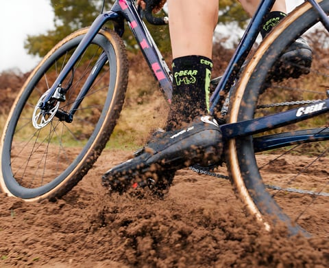 CX Syndicate rider kicks up soil on a navy cyclocross bike with tan-wall tyres and C-Bear socks.