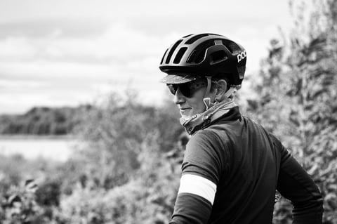 Black-and-white portrait of a rider in a POC helmet, cap and sunglasses glancing back beside a lake.