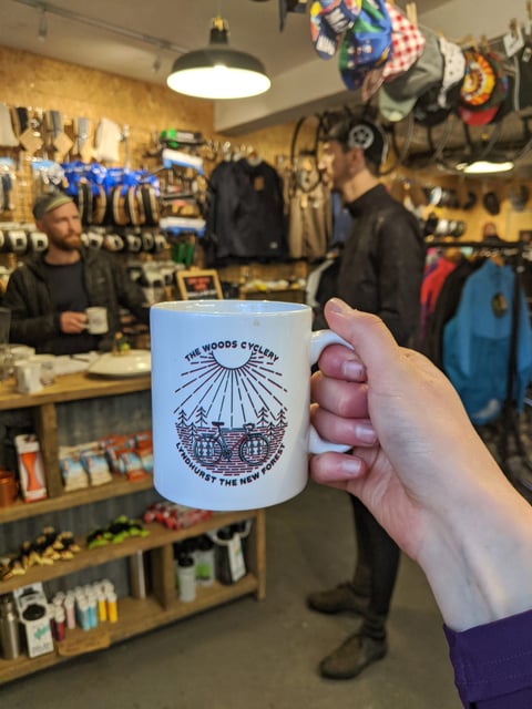 Hand holding a white The Woods Cyclery mug with a bike and sunburst design, inside the shop in Lyndhurst, the New Forest.