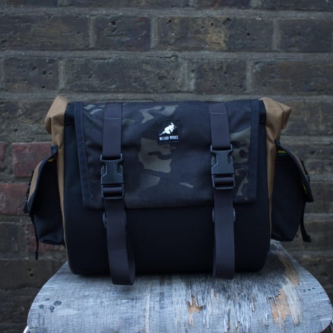 Wizard Works saddle bag in black with a camouflage flap and tan sides, on a stool in front of a brick wall.
