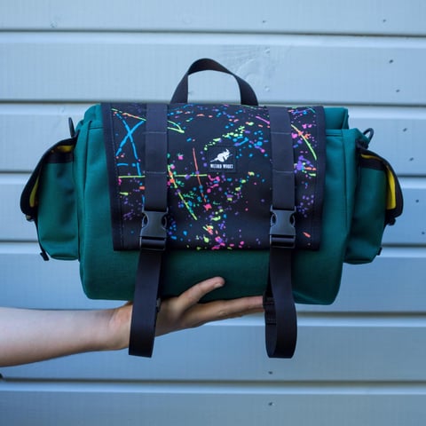 Wizard Works saddle bag in dark green with a neon splatter flap and yellow lining, held up against a pale wall.