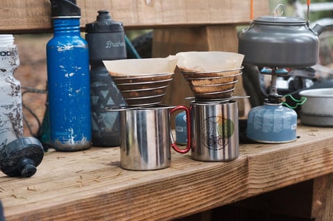 Two steel mugs with coffee filters, one printed with The Woods Cyclery logo, next to bottles and a camp kettle on a bench.