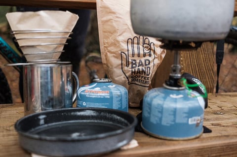 Camp stoves, filter cones, a steel mug and a Bad Hand Coffee Roasters paper bag on a wooden bench outdoors.